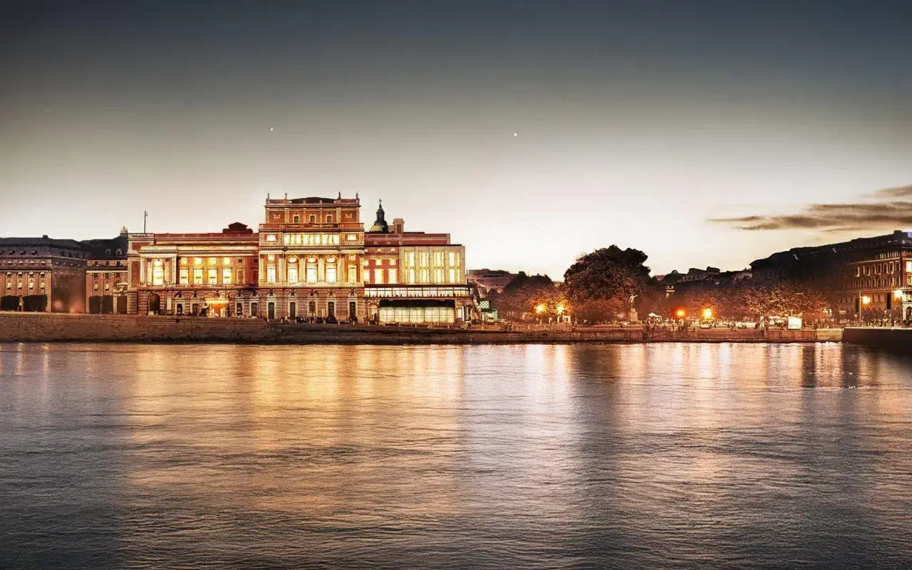 Kungliga operan i Stockholm lyser upp vid vattnet i skymningen, dess fasad speglas i det lugna vattnet.