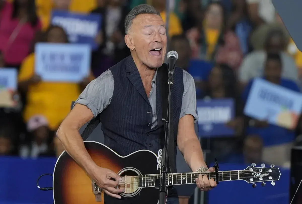 Bruce Springsteen sjunger och spelar gitarr på scenen. Publiken håller upp skyltar, vilket antyder ett kommande evenemang med Bruce Springsteen.