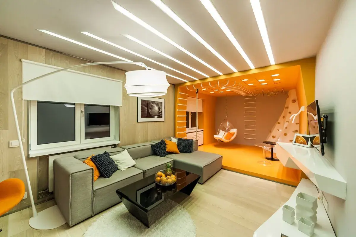 Stort vardagsrum med en grå soffa, svart soffbord och en orange sittgunga. Taket har en unik design med många ljuslister.