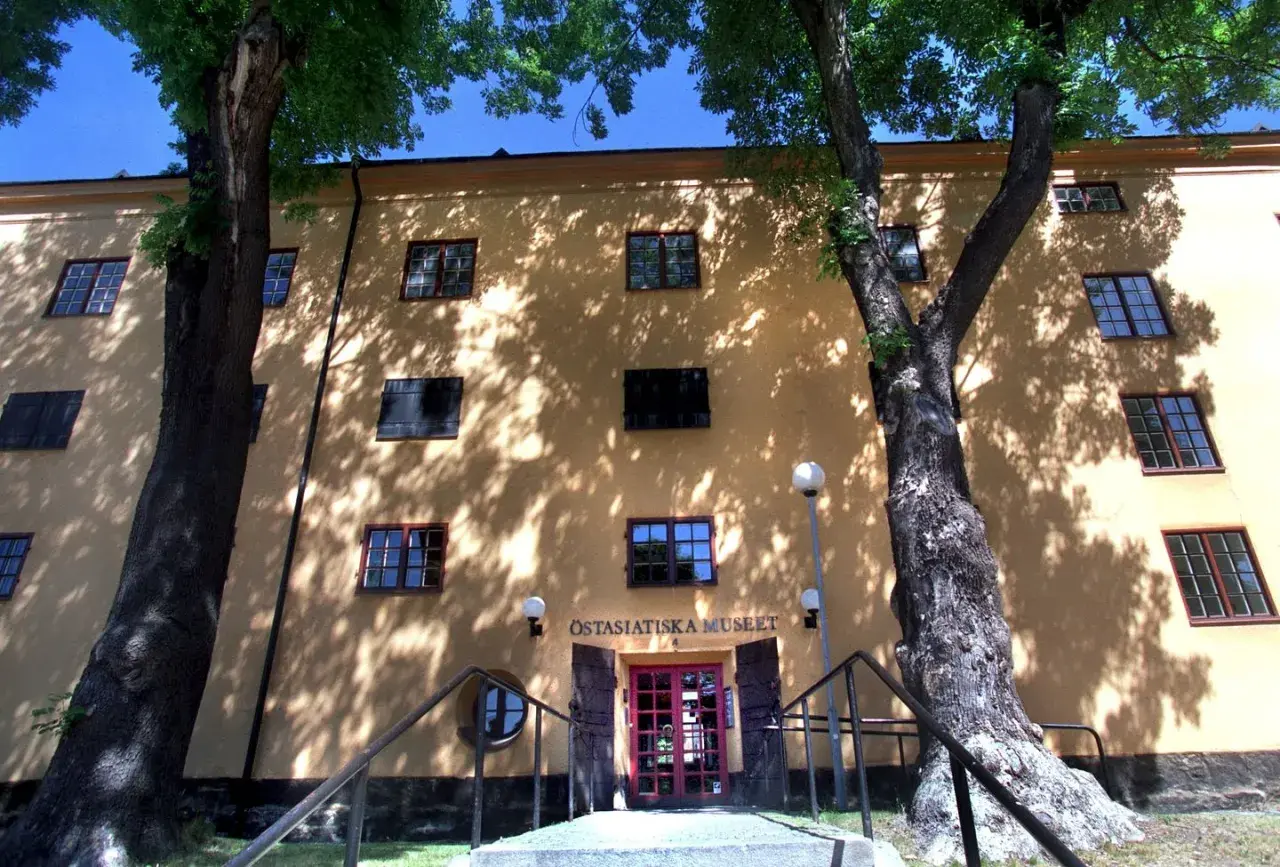 Östasiatiska museet i Stockholm, en gul byggnad med många fönster, omgiven av träd vars skuggor dansar på fasaden.