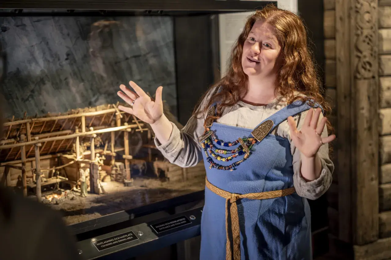 En kvinna i vikingakläder berättar vid en modell av en vikingagård på vikingamuseet.