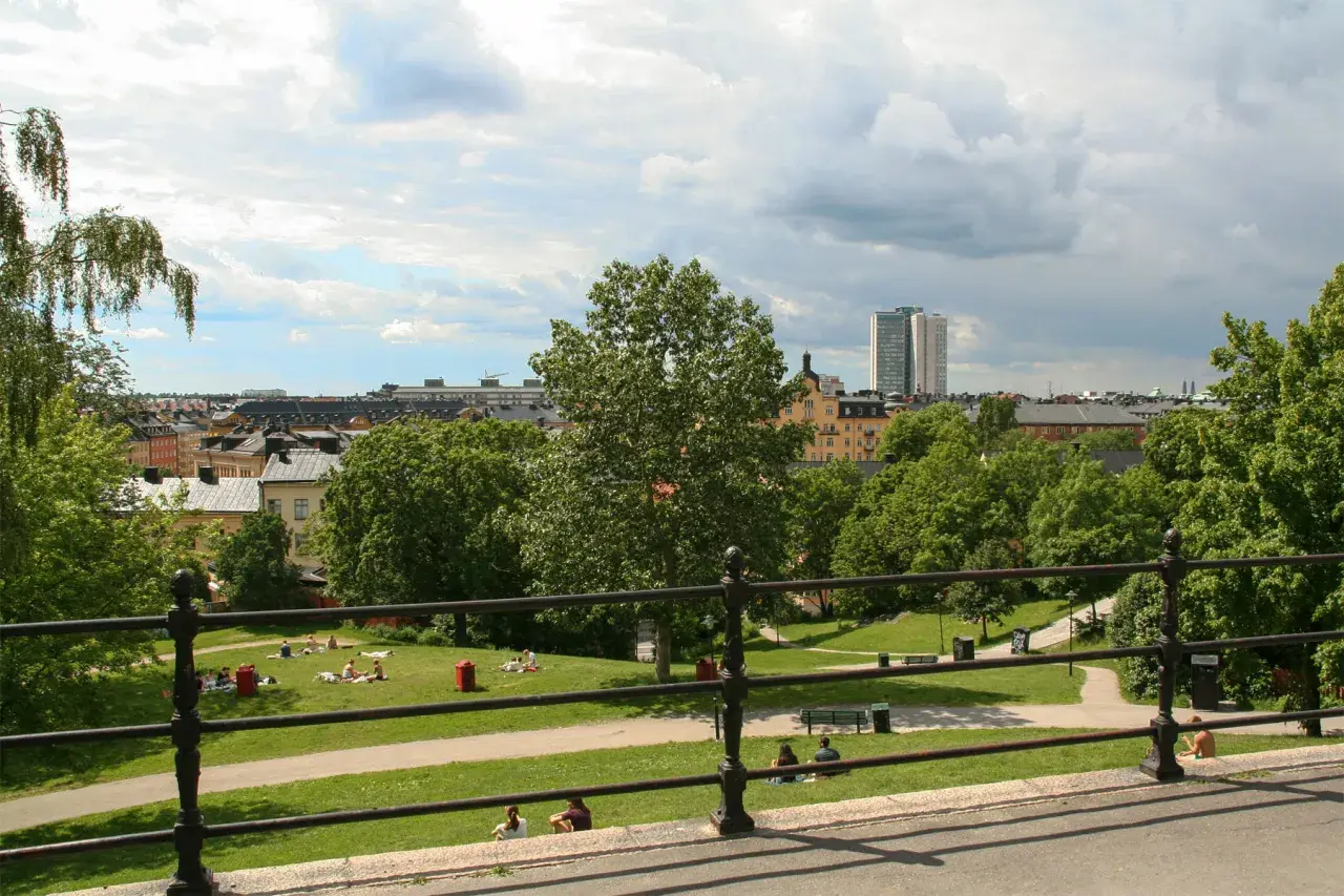 Sommar i Vitabergsparken, Stockholm. Människor njuter av solen på gräsmattan, med stadens takåsar och en teater i bakgrunden.