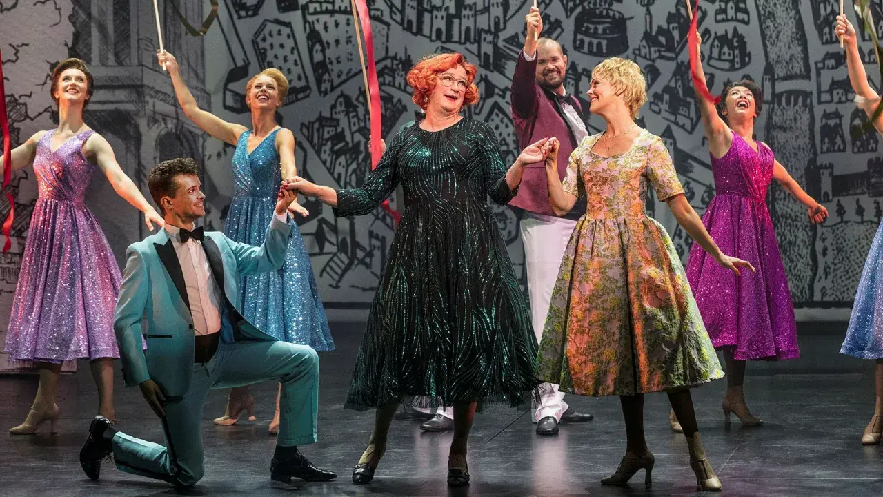 Scen från "Tootsie" på Oscarsteatern. En man i ljusblå kostym knäböjer medan en kvinna i glittrig grön klänning och andra i färgglada klänningar dansar.