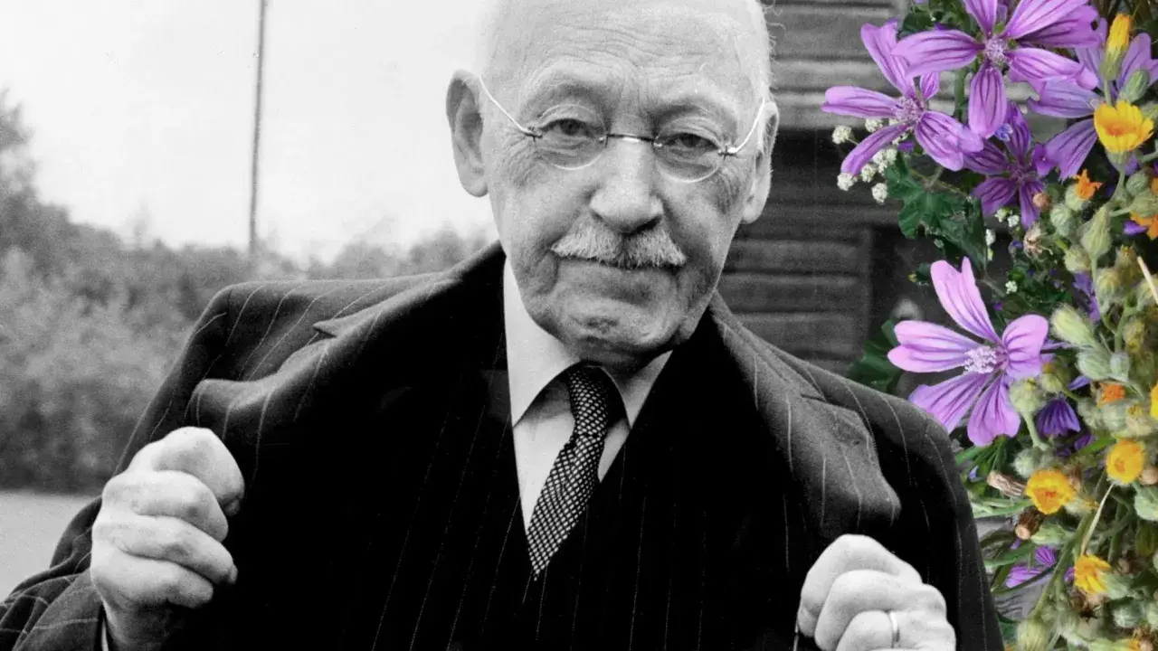 Äldre man med glasögon och mustasch, klädd i kostym. Bredvid honom en bukett med lila och gula blommor. Han är alfven som skrev.