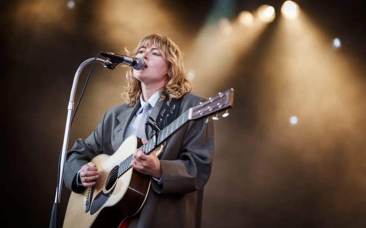 Ellen Krauss konsert. En artist med blont hår, klädd i kavaj och slips, spelar gitarr och sjunger i mikrofonen på en scen med strålkastare.