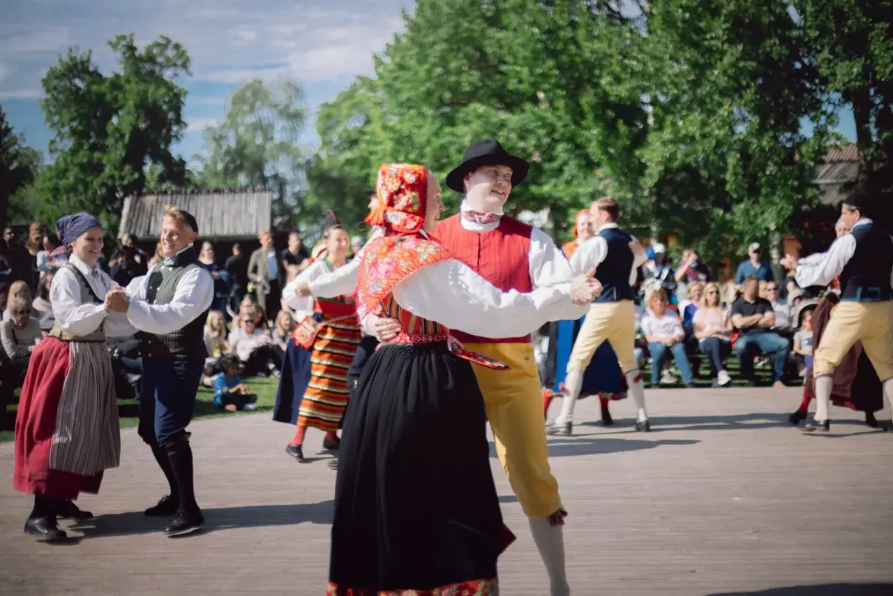 Dansande par i folkdräkter på en utescen, publiken tittar på. En härlig konsert på Skansen.