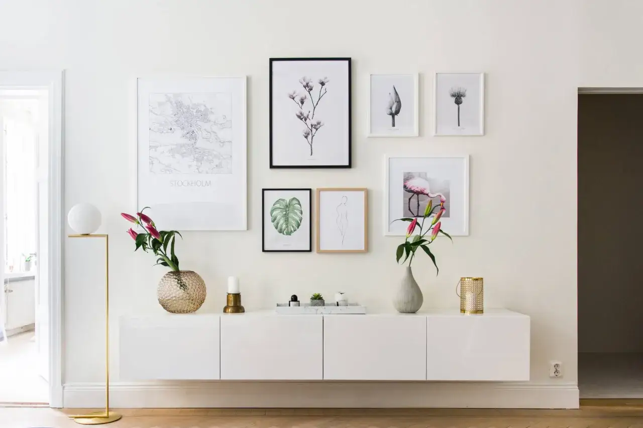 Ett modernt, vitt sideboard med konstverk ovanför, inklusive en Stockholmskarta och blomstermotiv. En golvlampa i mässing och en vas med blommor ger en stilfull touch.
