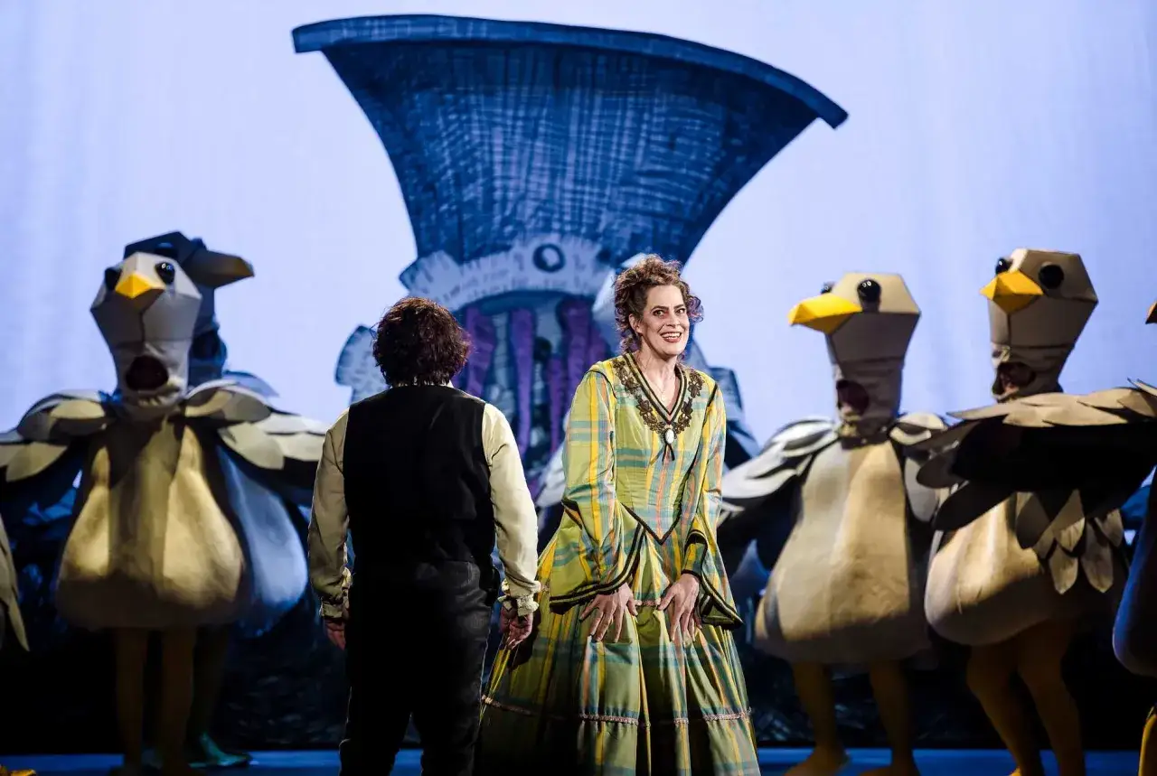 En kvinna i en rutig klänning ler mot publiken, omgiven av figurer i fågelkostymer på scenen för Sweeney Todd operan.