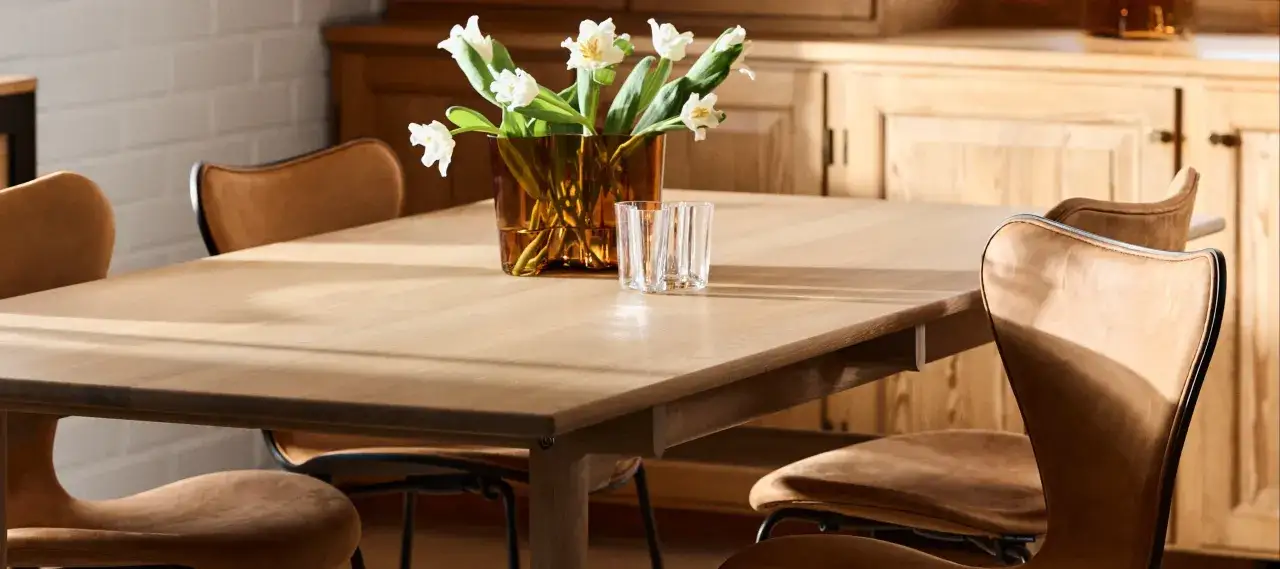 Ett ljust träbord med tulpaner i en bärnstensfärgad vas och glas. Stolar i ljusbrunt läder, typiskt för svenska designers.