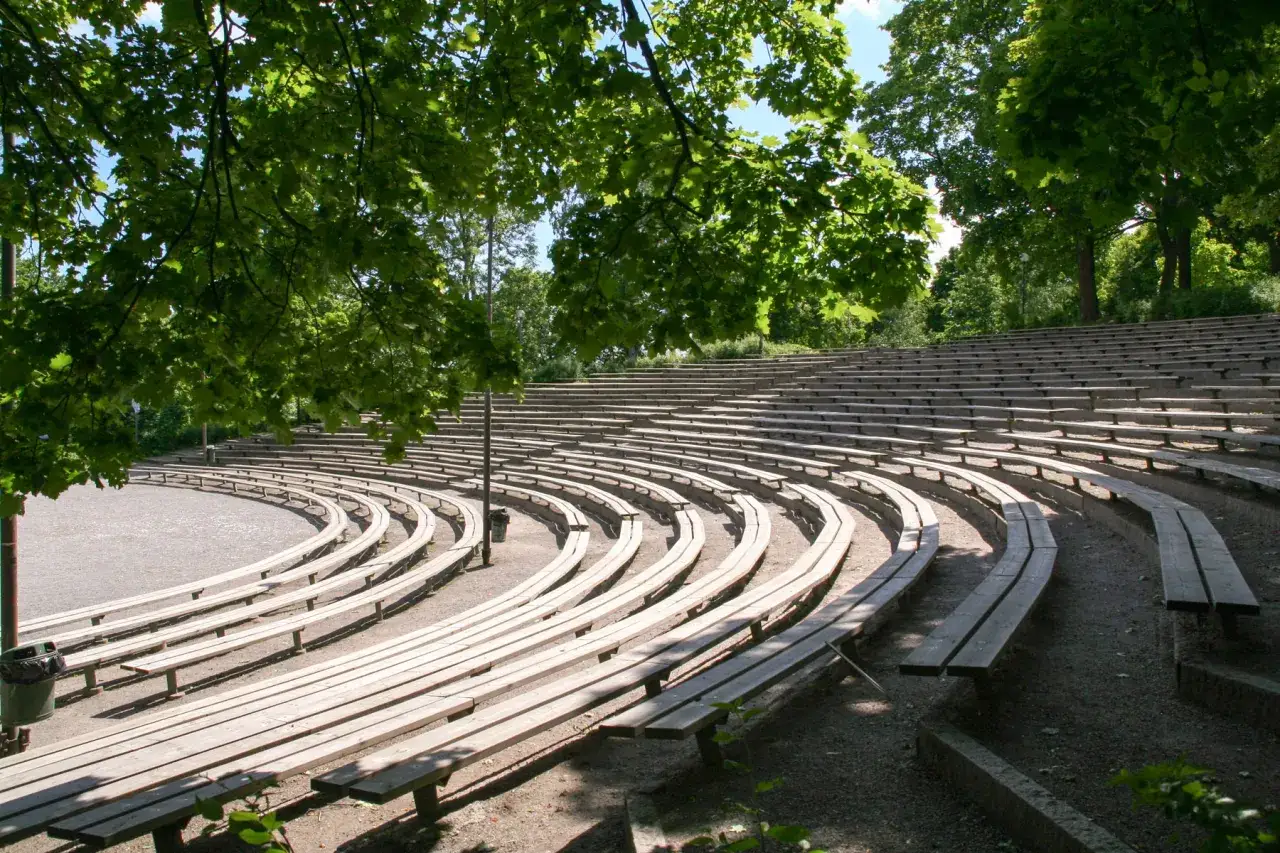 Tomma träbänkar i en cirkulär amfiteater i Vitabergsparken teater, omgivna av gröna träd.