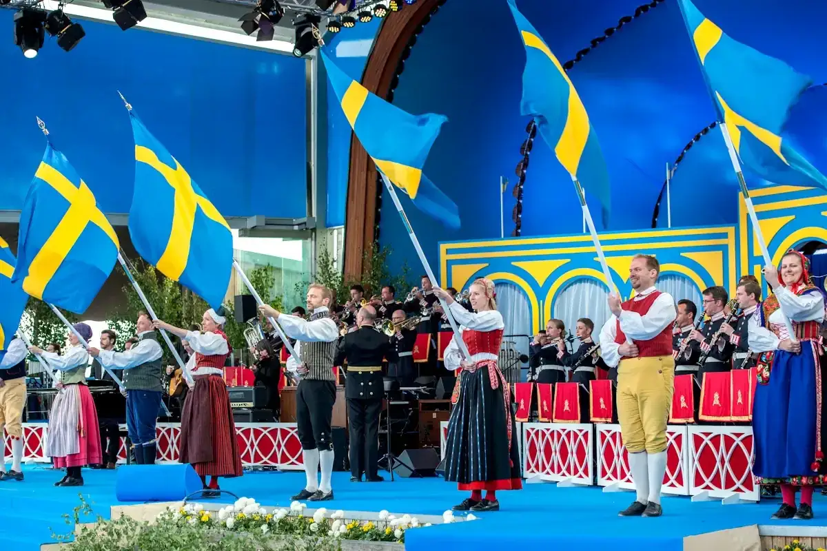 Människor i folkdräkter håller svenska flaggor under firandet av nationaldagen i Stockholm. En orkester spelar i bakgrunden.