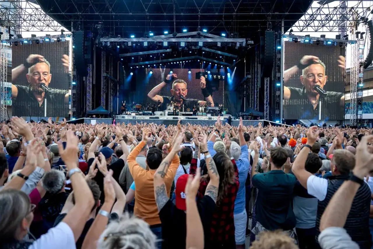 Publiken jublar vid ett kommande evenemang med Bruce Springsteen. Stora skärmar visar artisten på scenen.