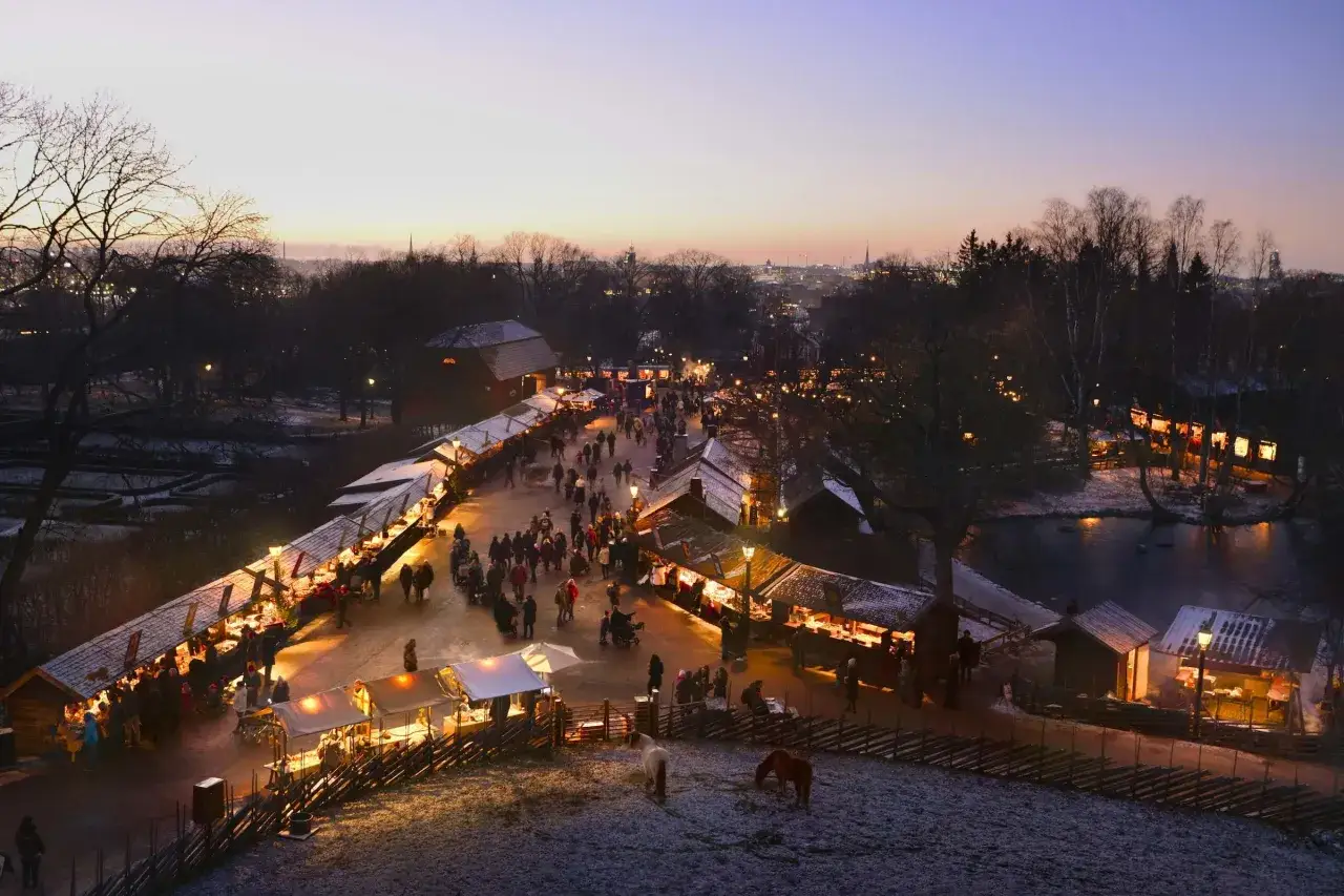 Vinterviken julmarknad i skymningen, med folk som strosar bland upplysta st&aring;nd och sn&ouml;kl&auml;dda tr&auml;d.