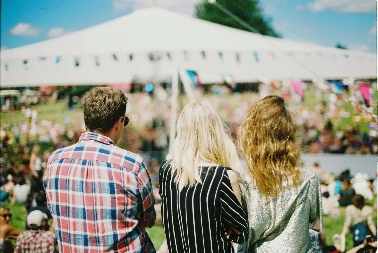 Tre vänner njuter av en solig dag på en av Stockholms festivaler. En stor vit tält och flaggor i bakgrunden skapar en festlig stämning.