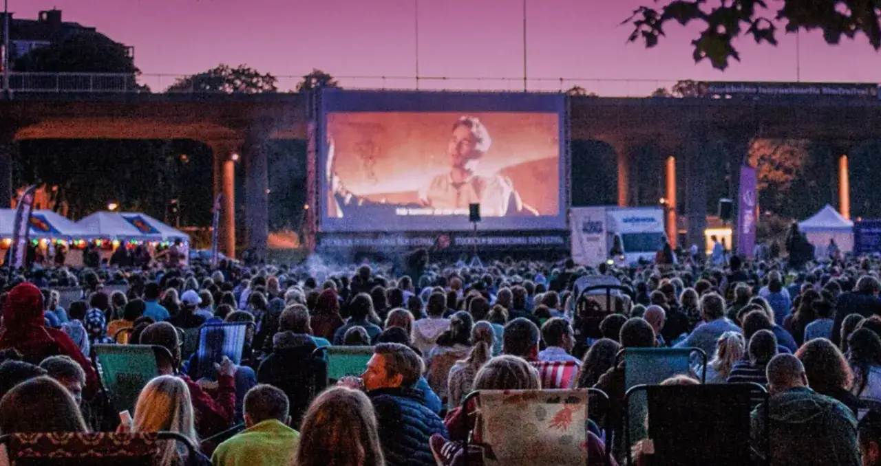 Stor publik njuter av utomhusbio i R&aring;lambshovsparken. En film visas p&aring; stor duk under en bro, med t&auml;lt och ljus i bakgrunden.