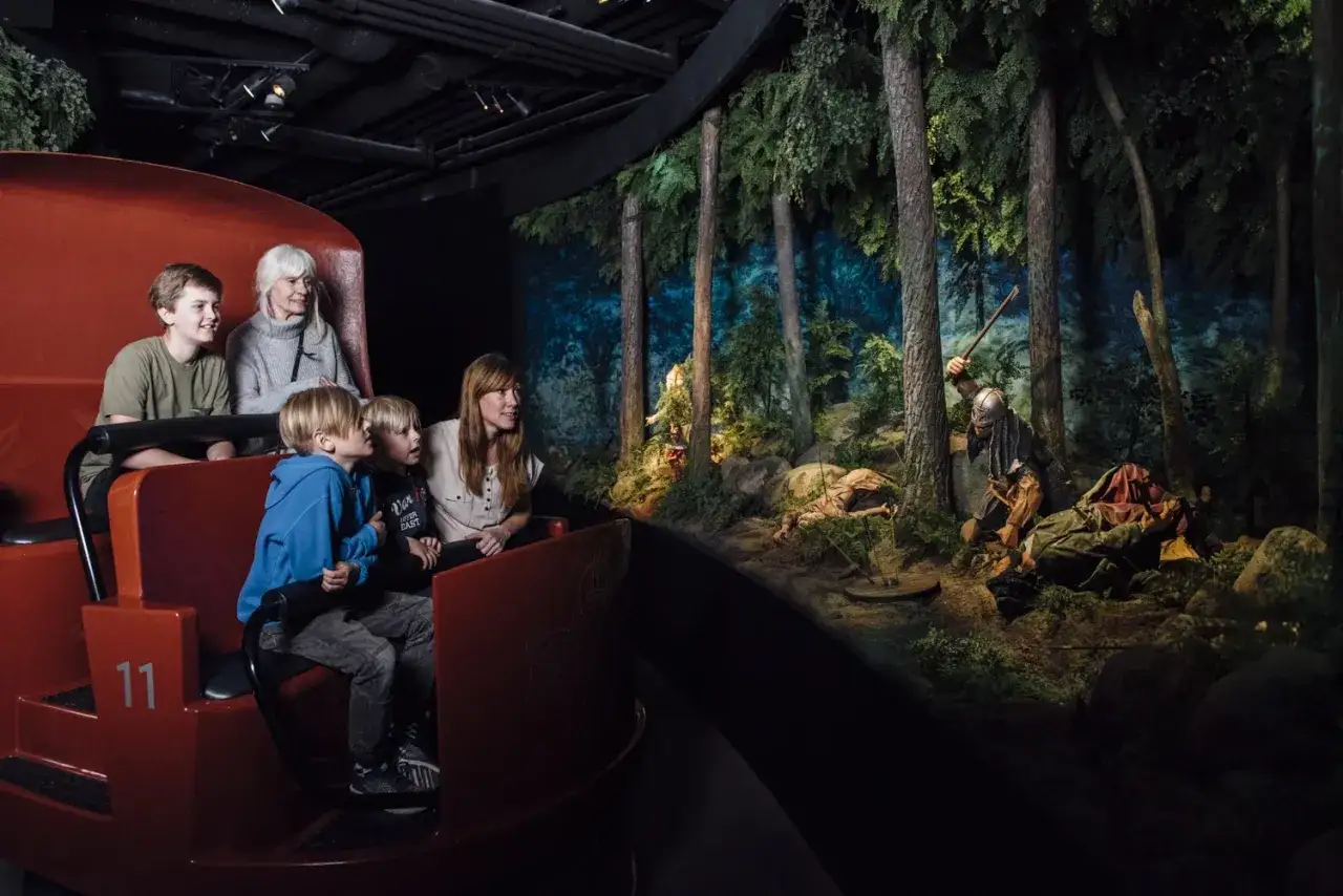 Familj i vagn på vikingamuseet ser en dramatisk scen med vikingar i skogen.