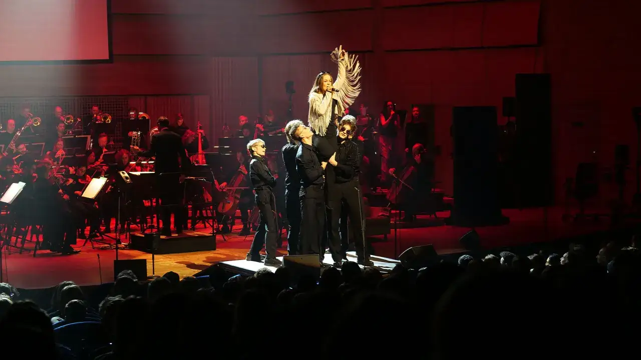 En ung artist sjunger p&aring; scenen under ett de geerhallen evenemang, omgiven av en orkester och publik.