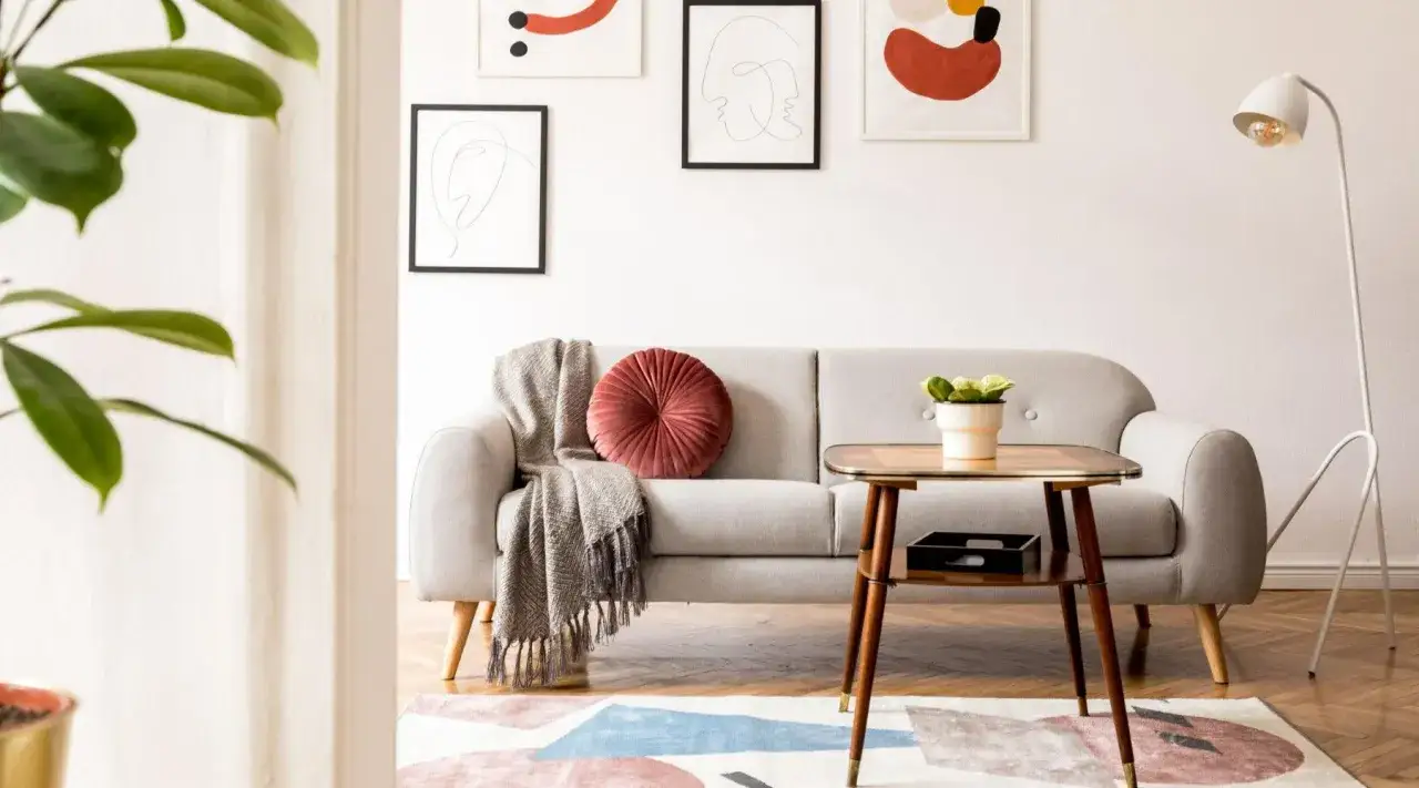 En ljusgrå soffa med en rosa kudde och pläd. Ett sidobord i teak med en krukväxt. Konst på väggen och en golvlampa ger en känsla av 50-tals inredning.