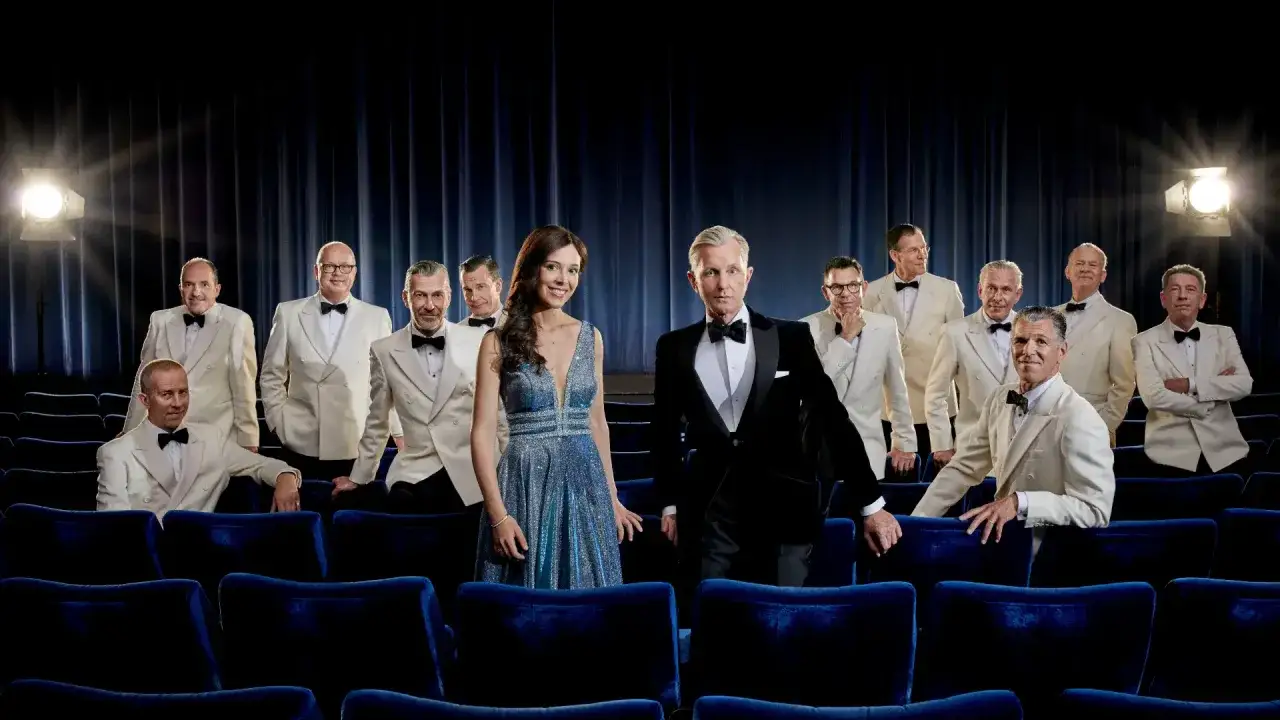 Max Raabe och hans Palast Orchester i Stockholm, klädda i eleganta vita kostymer och en glittrig blå klänning, poserar framför en mörkblå ridå.