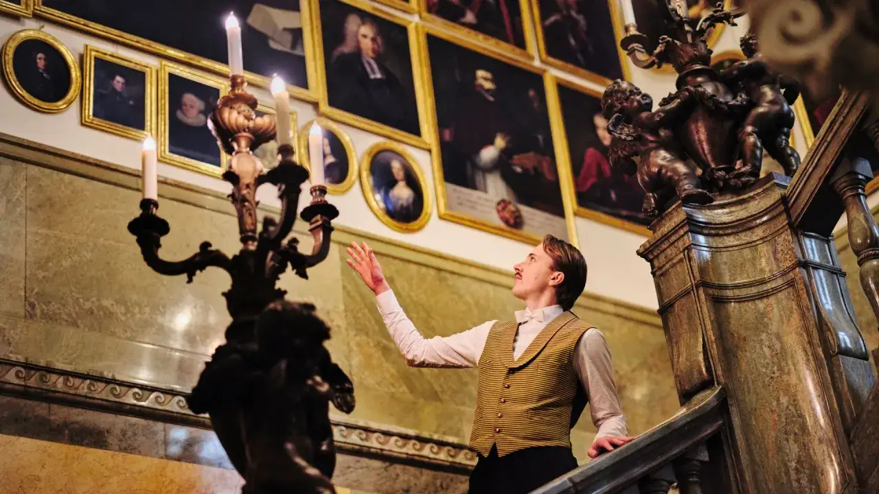 En man i historisk dräkt på trappan till Hallwylska museet i Stockholm, omgiven av porträtt och en ljusstake.
