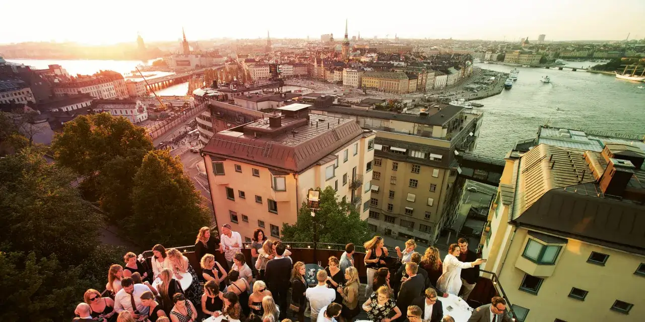 Festligt mingel på en takterrass med utsikt över Stockholm, kanske vid Mosebacke teater. Solnedgången färgar himlen gyllene.