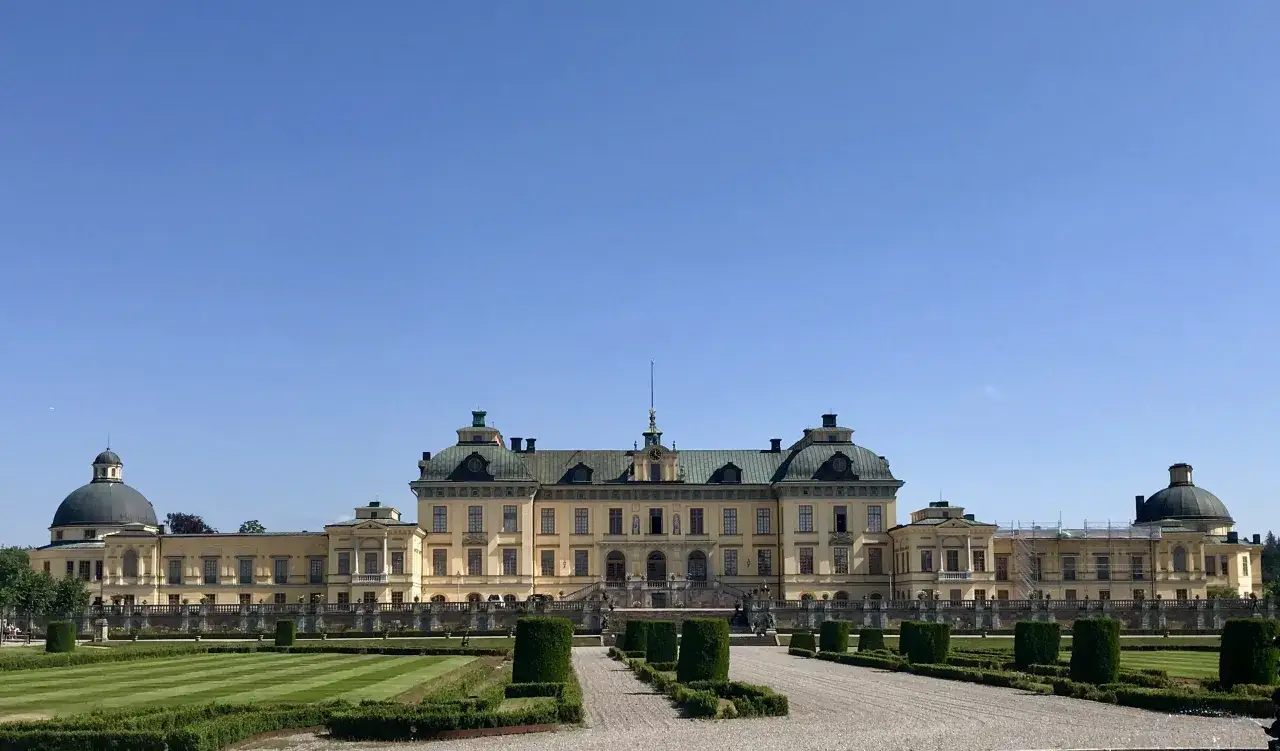 Drottningholm slott, ett ståtligt barockpalats med kupoler och symmetriska trädgårdar, badar i solljus under en klarblå himmel.