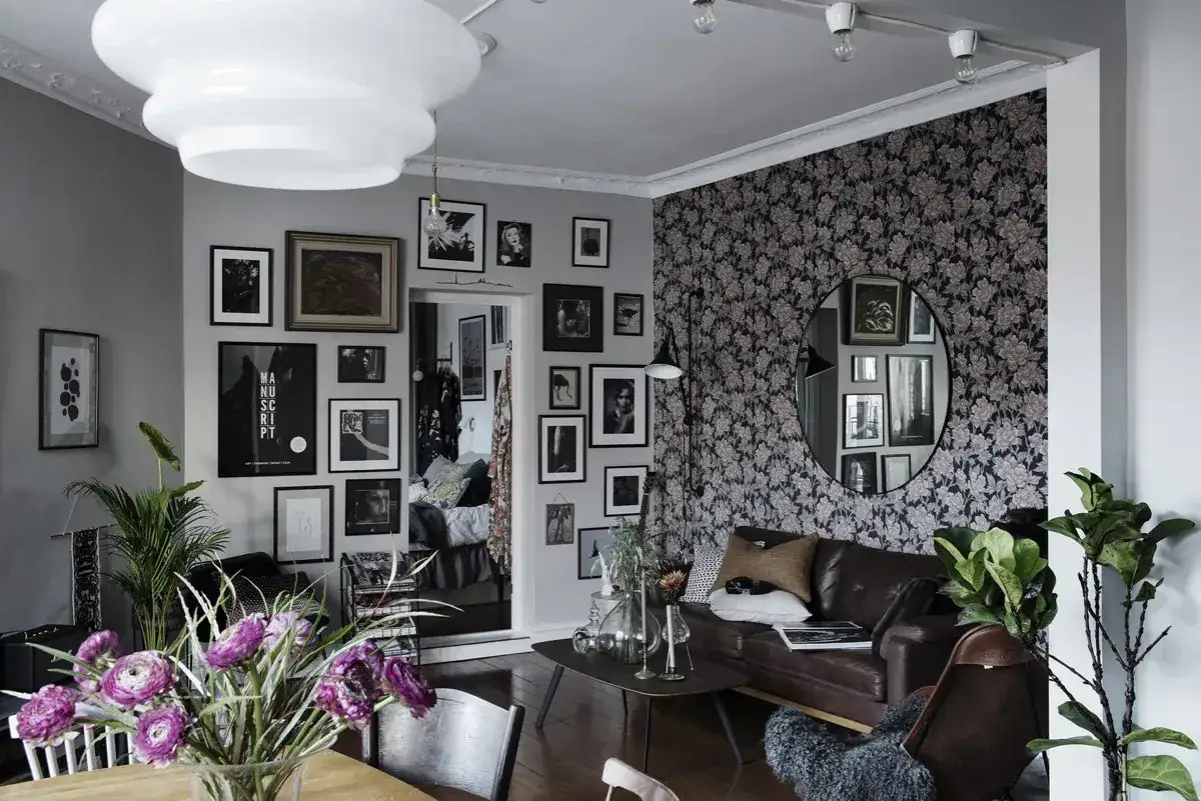 Ett vardagsrum i bohemisk stil med en vägg täckt av blommig tapet, en lädersoffa, många tavlor och en stor rund spegel.