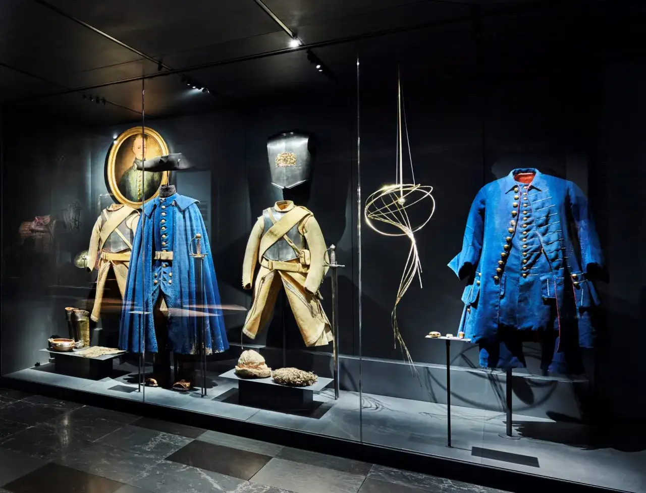 Kungliga Livrustkammaren visar historiska dr&auml;kter, rustningar och vapen. H&auml;r syns en bl&aring; uniform, en gul jacka med byxor, en br&ouml;stpl&aring;t och en portr&auml;ttm&aring;lning.