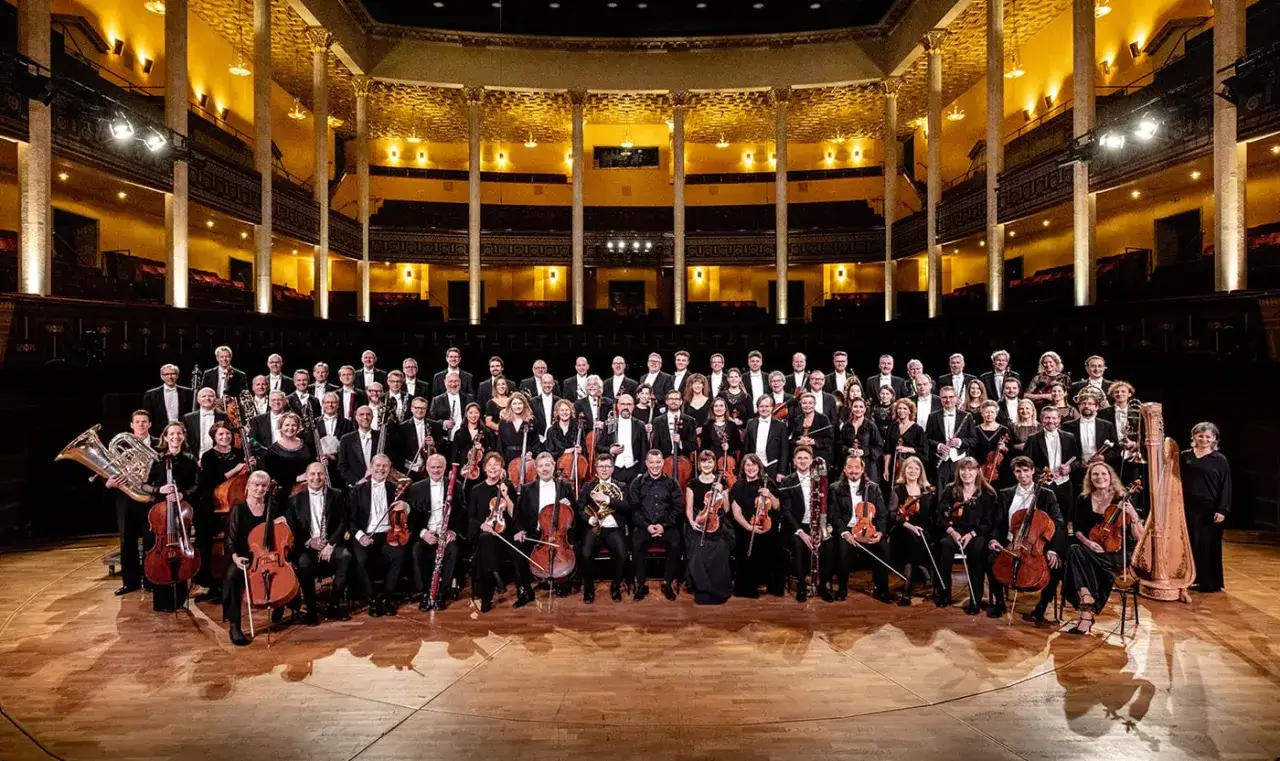 Kungliga Filharmonikerna medlemmar poserar i en stor orkestersal, redo att framföra musik.