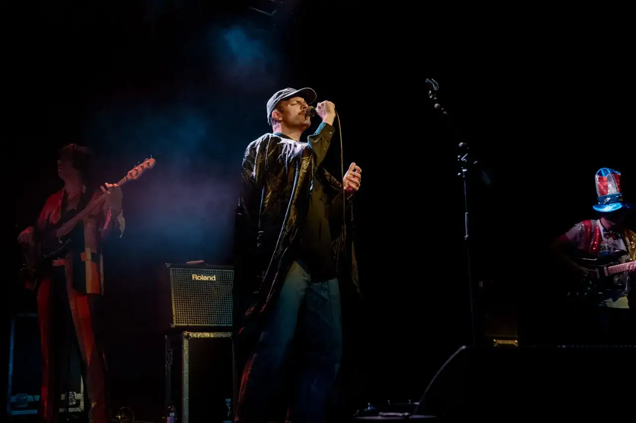 Mac DeMarco sjunger p&aring; scen i Stockholm, ikl&auml;dd keps och en glansig jacka. En basist och en gitarrist syns ocks&aring;.