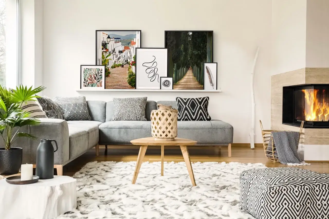 En modern soffgrupp med gr&aring; soffa och m&aring;nga kuddar. En hylla ovanf&ouml;r med flera tavlor som &auml;r fint placerade. En brasa sprakar i en &ouml;ppen spis.